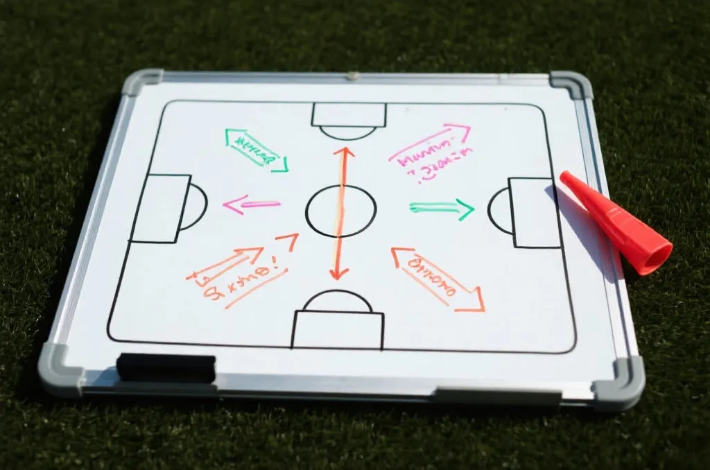 An overhead shot of a tactical whiteboard with soccer formations, arrows, and notes drawn in marker next to a whistle
